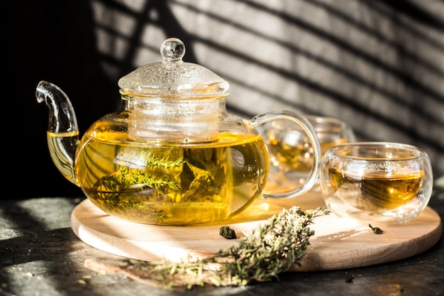 Yellow herbal tea in a transparent glass teapot and two bowls on a round wooden tray.