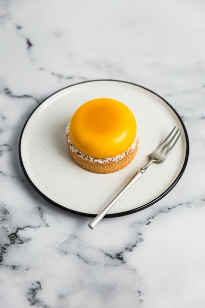 A yellow mango mousse tartelette on a white plate, a dessert fork,