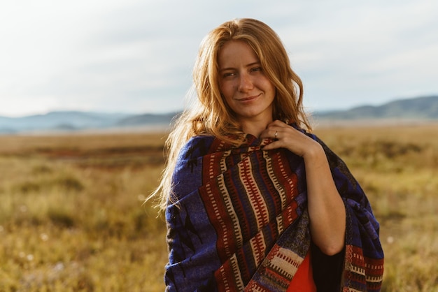 On the yellow meadow grass stands a girl of natural natural beauty with red hair in a colored cape o