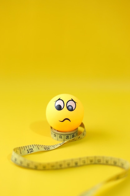 Yellow measuring tape with worried emoticon on yellow background Copy space for dietary program