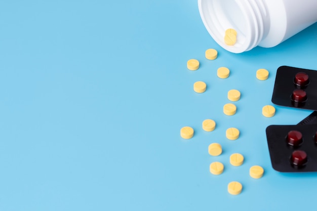 Yellow medical pills spilled out of white bottle and tablets on blue background.