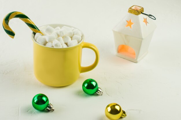 Yellow mug with Christmas hot cocoa and marshmallow, with caramel on a white background with glass balls and a burning candle.