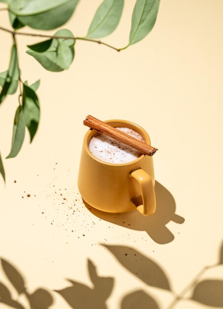 写真 黄色いカップ泡のある飲み物コーヒーラテホットチョコレートまたはココア硬い影と緑色の枝を持つ黄色い背景のシナモン棒