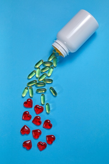 Photo yellow omega 3 gelatin tablets and red hearts spill out of a white jar on a blue background