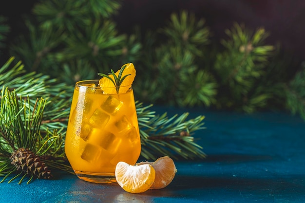 ダークブルーのコンクリート背景にガラスにタンジェリンとローズマリーを入れた黄色のオレンジ色のカクテルは、コーンで松の枝を飾り、クリスマスと新年のウェルカムドリンクを接写