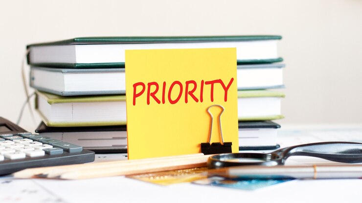 Yellow piece of paper with text priority stands on a clip for papers on the desk against books stacked, calculator, credit cards. Business and financial concept. Selective focus.