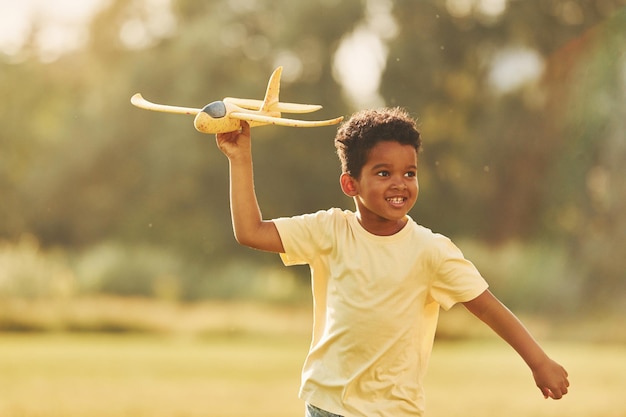 Yellow plane in hands African american kid have fun in the field at summer daytime