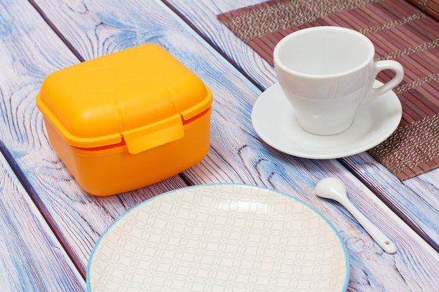 Yellow plastic lunch box, a white porcelain cup with saucer on a bamboo napkin, a plate on the wooden surface