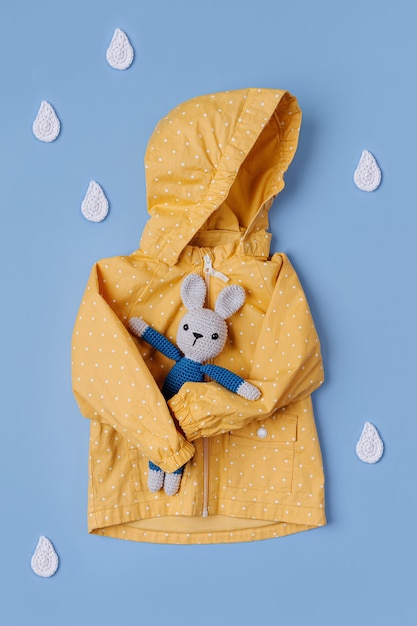 Yellow raincoat  with toy bunny and decorative raindrops on blue background. Autumn childrens outfit. Fall mood