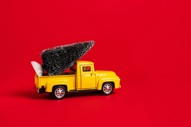 Yellow retro toy pickup carrying a Christmas tree on red background. Christmas celebration concept