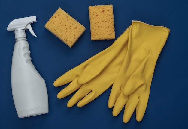 Yellow rubber gloves, sponges and bottle spray of detergent on\
classic blue background. color 2020. house cleaning theme. top\
view