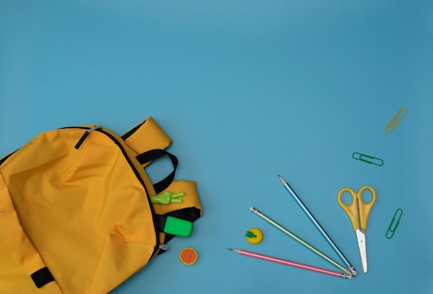 Yellow school knapsack with school or office supplies on blue background. back to school stationary