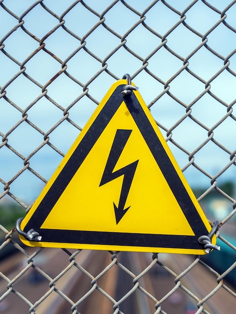 Фото Желтый знак с молнией в треугольнике знак висит на заборе цепной сетки