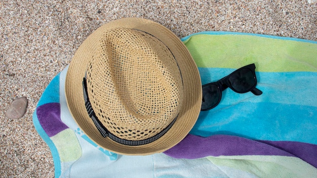 Yellow straw hat shade towel of blue and green colors on the white sand and next to sunglasses