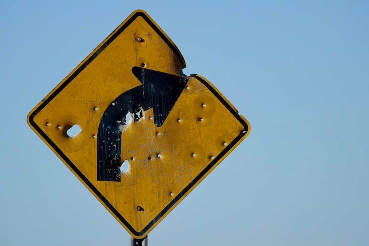 A yellow street sign indicating a curve in the road ahead is riddled with bullet holes