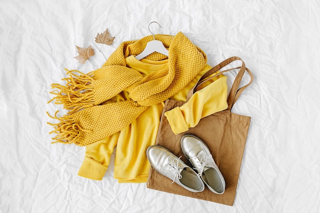 ニットスカーフ、靴、トートバッグ付きの黄色いセーター。白い背景の上の秋のファッションの服のコラージュ。上面図フラットレイ。