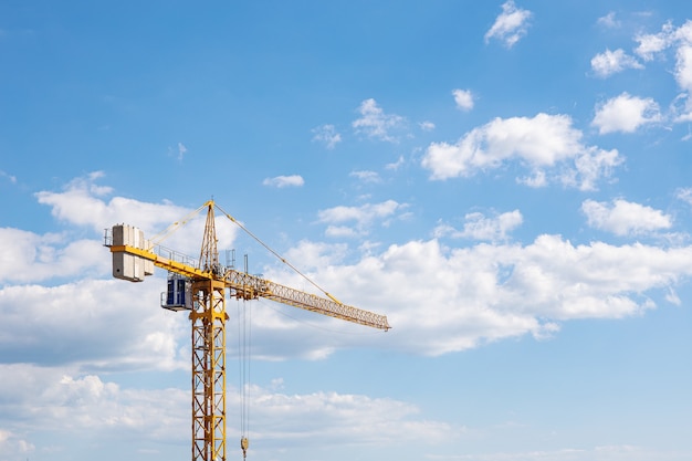 Yellow tall construction crane against the background of blue sky and clouds.