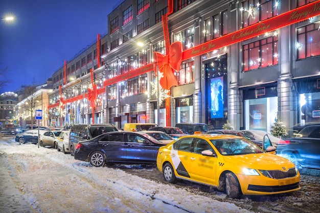 Taxi giallo e auto davanti al centro commerciale centrale di mosca e decorazioni sulla facciata del negozio a forma di fiocco rosso alla luce delle luci della sera didascalia: buon anno!