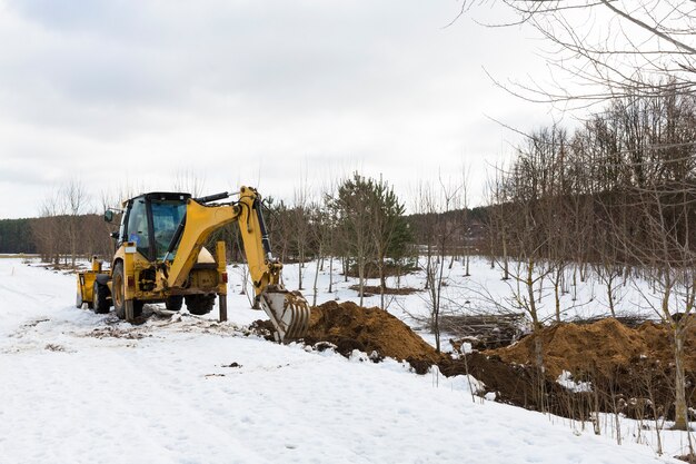 A yellow tractor excavator digs the ground for the repair and laying of underground utilities and cables for electricity and communications. During the winter season. High quality photo