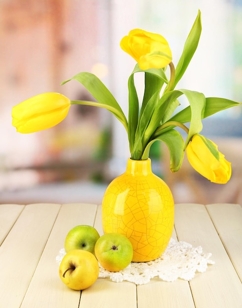 Yellow tulips in vase on wooden table on room background