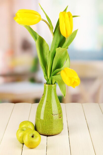 Yellow tulips in vase on wooden table on room background