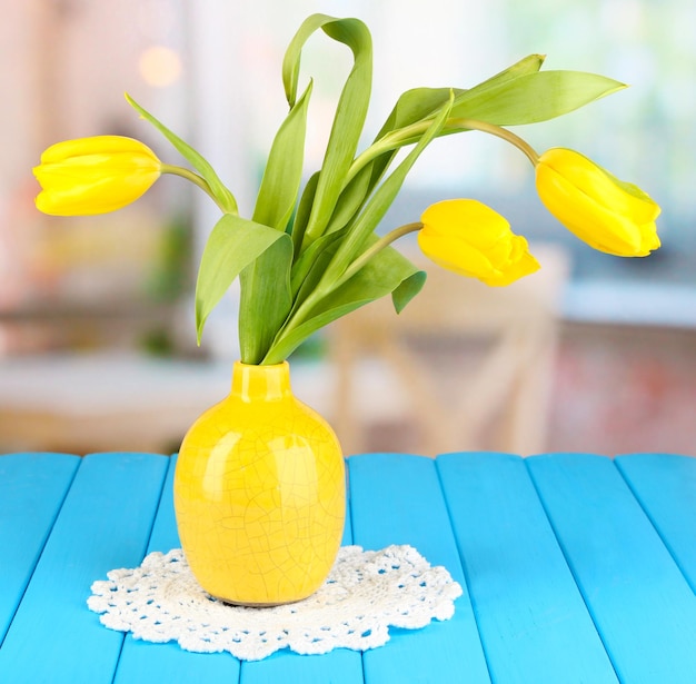 Yellow tulips in vase on wooden table on room background