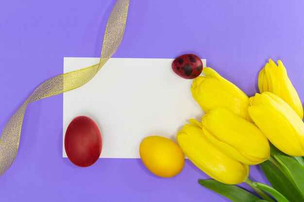 Желтые тюльпаны с пустым белым листом для текста или макета и окрашенные желтые и красные яйца золотая лента Пасхальный фон Копирование пространства