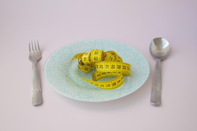 A yellow twisted centimeter ribbon lies in a blue plate next to a fork and spoon on a pink background