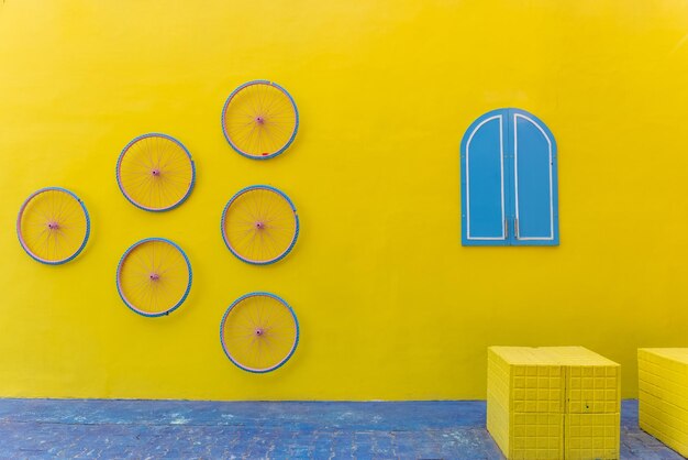 A yellow wall with four wheels on it and a window with blue shutters.