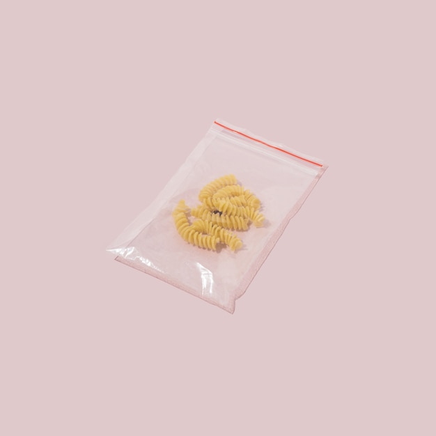 Желтые цельнозерновые спирали в небольшом полиэтиленовом пакете на пастельно-розовом фоне. Минимальный стиль концепции еды.