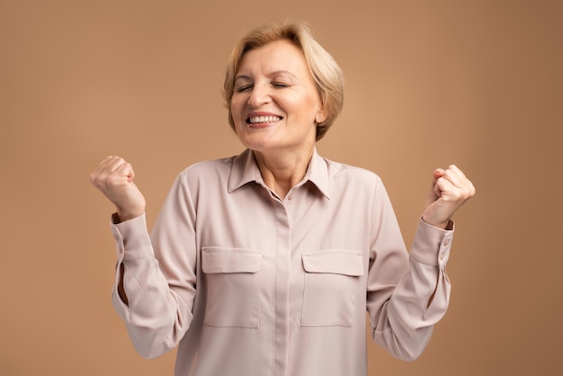 Yes. Extremely satisfied woman in casual clothes celebrating her victory rejoicing and showing yes gesture, positive emotions. Indoor studio shot isolated on brown background