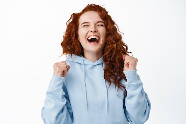 Yes hooray. Excited redhead girl winning and celebrating, shouting from satisfaction, making fist pumps and laughing, triumphing, achieve goal on white