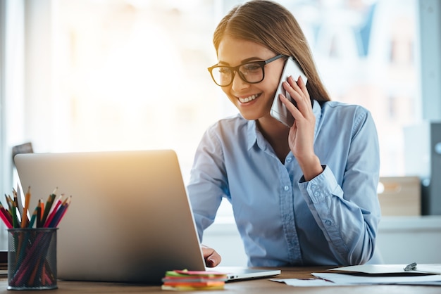 写真 はい、あなたのメールが表示されます。彼女の職場に座っている間、携帯電話で話し、笑顔でラップトップを使用してメガネで陽気な若い美しい女性