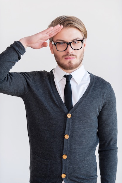 Photo yes sir! handsome young man in glasses holding hand near head and looking at camera while standing against grey background