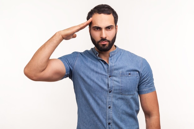 yes sir subordinate disciplined man with beard saluting holding hand near head looking at camera with serious expression patriotism and discipline indoor studio shot isolated on white background