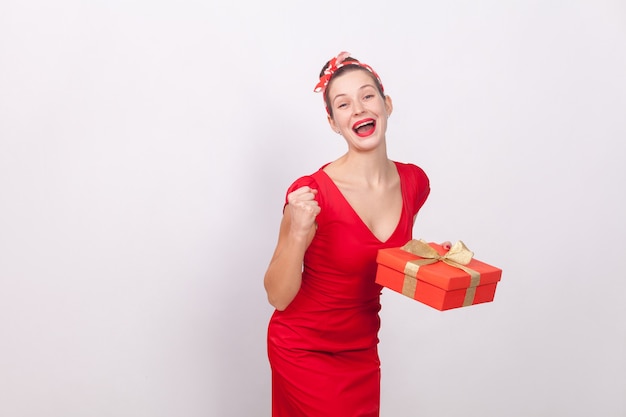 Yes! Win! Success beautiful woman holding gift box, toothy smile. Indoor, studio shot, isolated on gray background