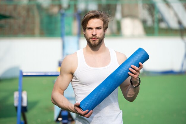 Yoga beoefenen. Sportman draagt yogamat om buiten te oefenen. Buiten yoga-concept. Man atleet zelfverzekerd gezicht draagt mat, gaat uitrekken. Atleet rekt zich altijd uit na de training.