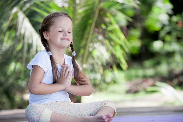 Ребенок йоги упражнения на деревянной платформе среди зеленых растений на открытом воздухе.