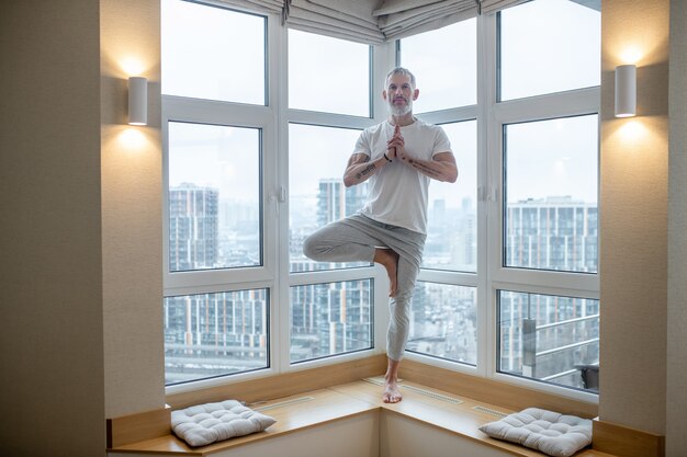 Yoga. Gray-haired man in a white tshirt standing in a tree pose