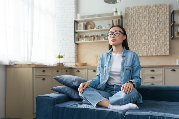 Yoga at home a young asian woman in glasses sits calm and
relaxed at home on the sofa in the lotus