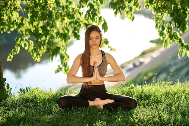 Yoga in het park met uitzicht op de rivier, met zonlicht. Jonge vrouw in lotus positie zittend op het groene gras. Het concept van rust en meditatie.