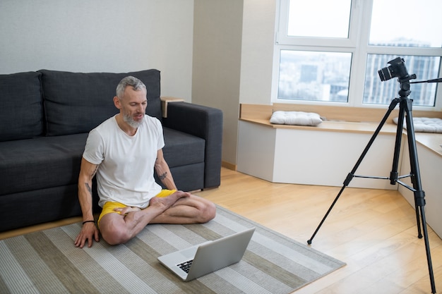 Yoga lessen. Een volwassen man in een wit t-shirt die op de grond zit en een online yoga-tutorial opneemt