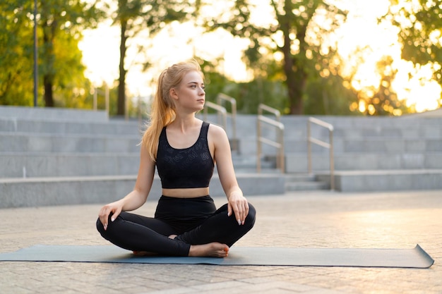 Yoga and Meditation in Modern City Caucasian Woman Relax Lotus Position Sitting Yoga Mat Outdoors Summer Park on Concrete floor Young Adult Fit Female Relaxing after Yoga Practice Workout Siddhasana