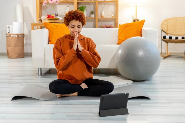 Yoga mindfulness meditation Young healthy african girl practicing yoga at home Woman sitting in lotus pose on yoga mat eyes close hands in prayer meditating indoor Girl doing breathing practice