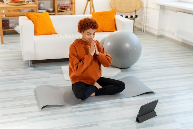 Yoga mindfulness meditation Young healthy african girl practicing yoga at home Woman sitting in lotus pose on yoga mat eyes close hands in prayer meditating indoor Girl doing breathing practice