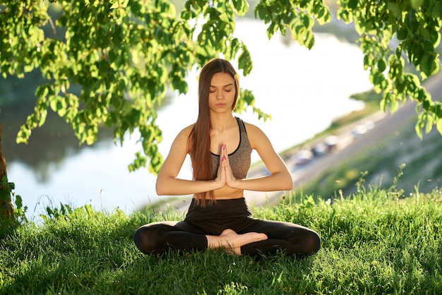Йога в парке с видом на реку, с солнечным светом. Молодая женщина в позе лотоса, сидя на зеленой траве. Понятие спокойствия и медитации.