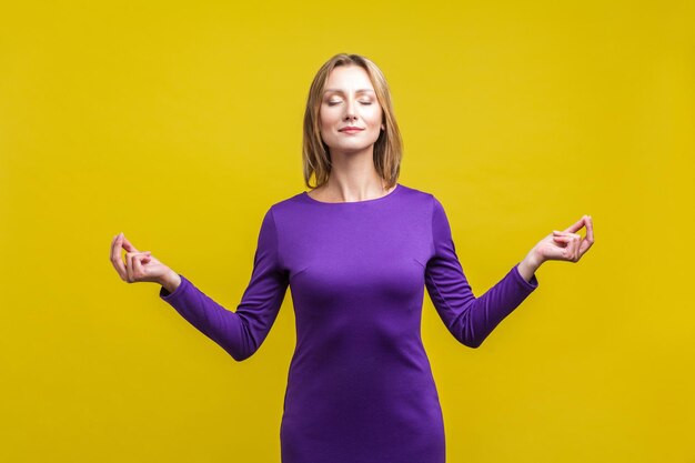 Yoga praktijk harmonie portret van vreedzame vrouw in paarse jurk permanent met gesloten ogen en kalm gezicht mediteren met vingers in mudra gebaar studio opname geïsoleerd op gele achtergrond