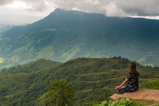 Женщина йоги сидя делающ раздумье на утесе с предпосылкой горы.