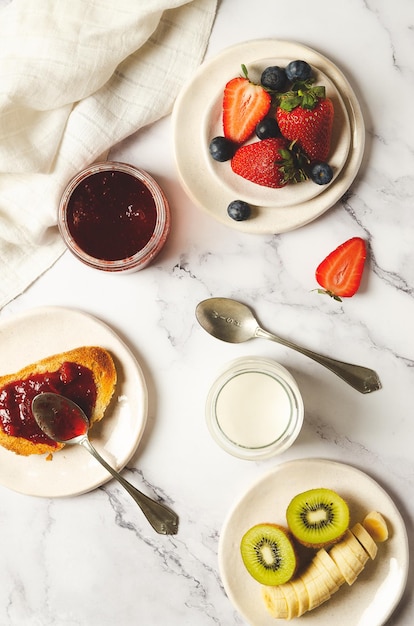 Yoghurt, fresh fruits, strawberry jam, and bread with a white napkin on marble background.