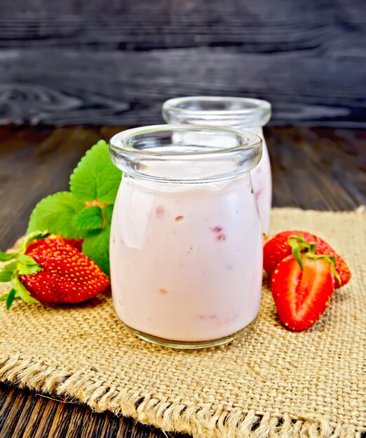 Yoghurt met aardbeien in twee glazen pot op een servet van jute met bessen en munt op een houten planken achtergrond